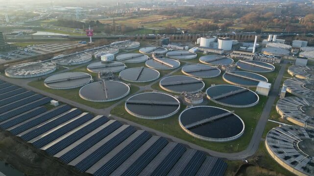 Smooth cinematic drone movement above large circular sewage clarification tanks at the Harnaschpolder wastewater treatment plant in the Netherlands surrounded by green infrastructure.