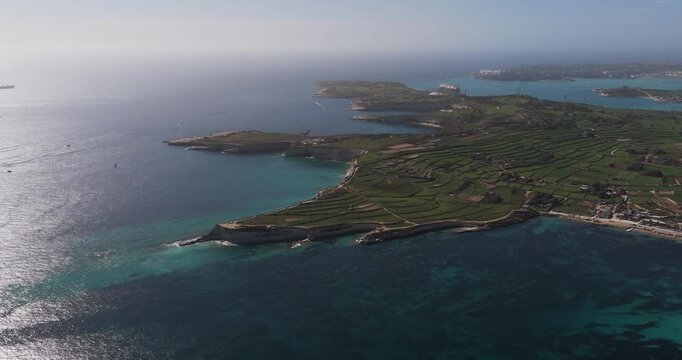 Aerial view of the rugged coastline meeting the clear turquoise waters, contrasted by the lush green fields inland, Munxar Path, Marsaskala, Malta.