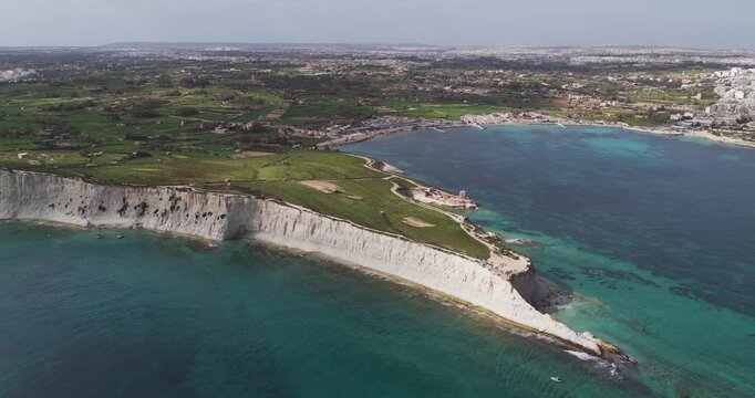 Aerial view of the white cliffs meeting the turquoise sea near Munxar Path, with green fields stretching inland, Marsaskala, Malta.