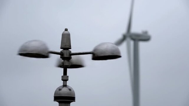 A close-up shows a spinning cup anemometer measuring wind speed against a blurry backdrop of rotating wind turbines under an overcast, grey sky, focusing on renewable energy and weather data collecti