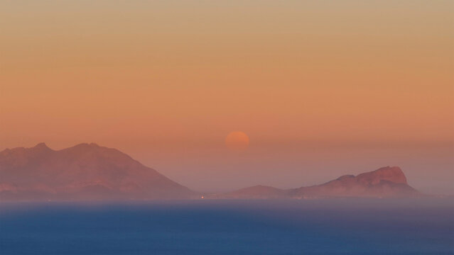 Aerial view of mountains shrouded in a soft, warm haze, with the sun appearing as a fiery orb on the horizon, Cape Town, Western Cape, South Africa.