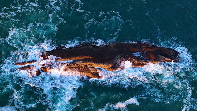 Aerial view of a rugged island kissed by golden light amid the swirling, frothy turquoise waters of the ocean, Cape Town, Western Cape, South Africa.