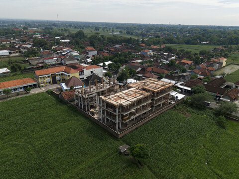 Aerial view of a building under construction, surrounded by lush green fields contrasting with the dense residential area, Klaten Regency, Central Java, Indonesia.