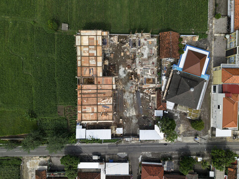 Aerial view of a building under construction juxtaposed against the verdant rice paddies and the contrasting red-tiled roofs of adjacent houses, Klaten Regency, Central Java, Indonesia.