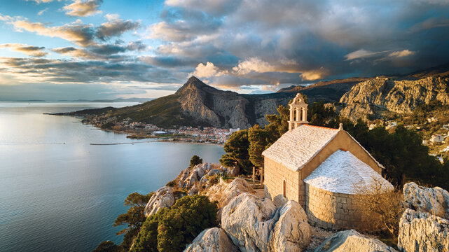 Aerial view of a quaint church perched atop rocky cliffs overlooking the Adriatic Sea, embraced by the dramatic Dinaric Alps under a sky ablaze with golden light, Omis, Split-Dalmatia County, Croatia.