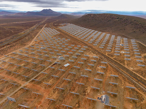 Aerial view of a vast solar farm shimmering under the diffused light, contrasted against the arid landscape and distant mountains, Touws River, Western Cape, South Africa.