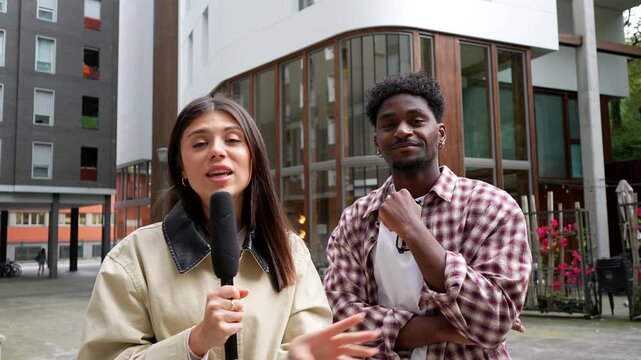 Young female reporter with a microphone conducting a street interview with an african american man in a modern urban setting, both smiling and waving at the camera at the end of the take