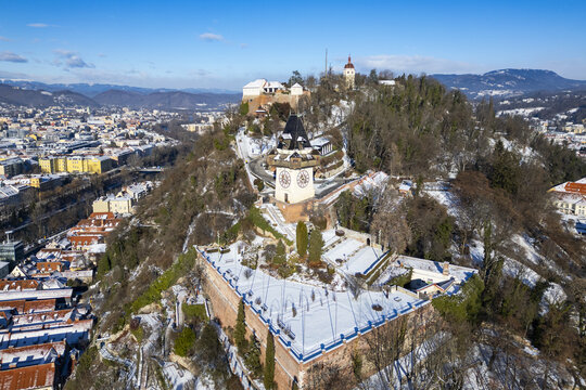 Aerial view of the Uhrturm clock tower and the Grazer Schlossberg stand majestically against a backdrop of snow-dusted rooftops and clear blue skies, Graz, Styria, Austria.
