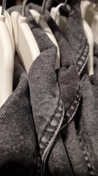 Dark grey denim shirts hang in a row on white plastic hangers on a clothing rack. A close-up, handheld shot focuses on the texture of the fabric and the collar stitching.