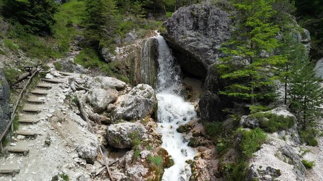Upward drone glide follows Ciastlins waterfall channel near San Vigilio Marebbe, multiple streams tumble through granite cleft beside trail steps and alpine larch