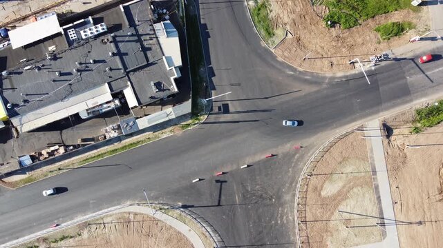 Top-down aerial shot of an unregulated road intersection undergoing construction. New asphalt, dirt patches, and sparse traffic show urban infrastructure development in progress. Dynamic scene