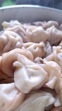 Close-up view of freshly cooked pelmeni dumplings piled in a white bowl. Hot steam rises from the moist dough pockets filled with meat, highlighting the soft appetizing texture and heat.