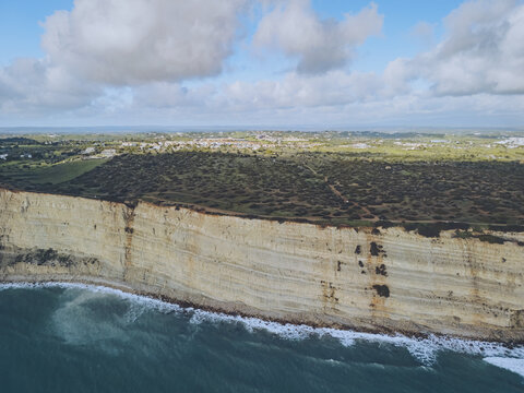Aerial view of the rugged, layered cliffs meeting the turquoise sea, contrasting with the lush greenery above, Praia do Porto de Mos, Lagos, Faro, Portugal.