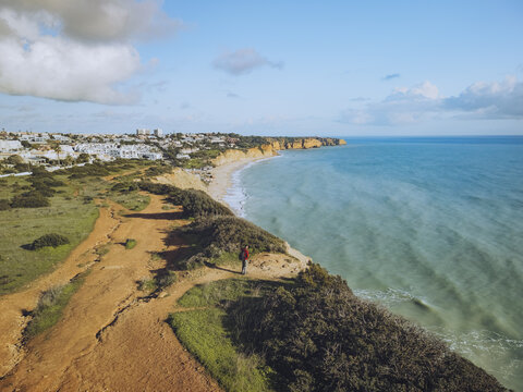 Aerial view of the rugged coastline and sandy beach kissed by the turquoise sea, with a lone figure admiring the vista at Praia do Porto de Mos, Lagos, Faro, Portugal.