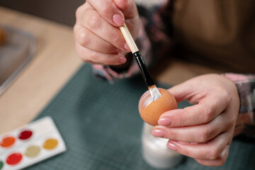 Easter eggs at home. Close-up of woman decorating painting easter egg using brush. Cozy atmosphere for holiday creativity and festive preparation.