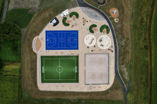 Aerial view of vibrant blue basketball court, a green soccer field, and a sandy volleyball court amidst lush greenery, Monthey, Valais, Switzerland.