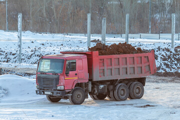 A red dump truck loaded with soil at a construction site in winter