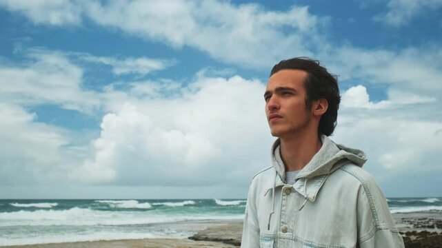 Young handsome man walks alone on the empty ocean beach on the cloudy day. Pretty guy walks a sandy shore and relaxing. Young Brunet man is walking against blue sea with clouds on the sky. Slow motion