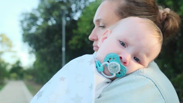 Baby rests on his mother's shoulder, in the park. Close-up of a calm and looking away little child on mom, outdoors. Child is resting  in the fresh air. Maternity  concept. Happy Baby sucks a pacifier