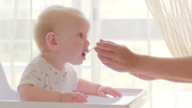 Slow motion: feeding a baby from a spoon. Mom feeds a 9 month baby with fruit puree from a spoon, close-up, high key. Meal time. Little boy eats sitting on baby's chair. Mother cares about son
