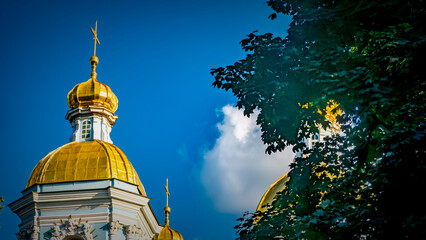 Golden domes of an Orthodox church against a vibrant blue sky. Lush green trees frame the architecture. A beautiful religious landmark