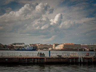 Historic city buildings line a waterfront under a dramatic sky filled with puffy clouds. A dark pier foreground adds depth to this European scene.