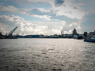 Dramatic river view with city skyline under stormy clouds. A pier extends into the water, featuring a crane boats. Moody lighting creates a striking scene