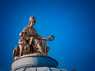 A striking low-angle shot of a bronze statue atop a dome against a vibrant blue sky. The sculpture depicts a regal figure with cherubs, radiating historical grandeur