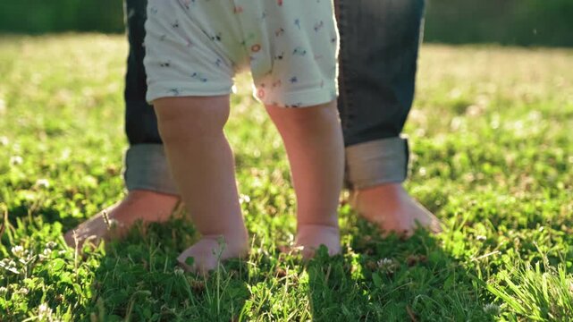 Walking children's bare feet on a green lawn close-up. Child  learns to take the first steps.  Baby learns to walk with the help of his mother on a green grass in the park. Slow motion 4k video.