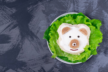 Bear cub, sandwich for kids with wheat bread and cheese, served on salad leaves on the gray table. Copy space. Top view © kcuxen