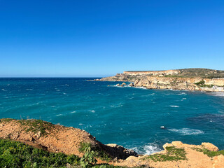 Fototapeta premium Wide elevated views of the north-west coast of Malta showing rugged limestone cliffs, eroded rock formations and a small sandy bay with turquoise Mediterranean Sea. 