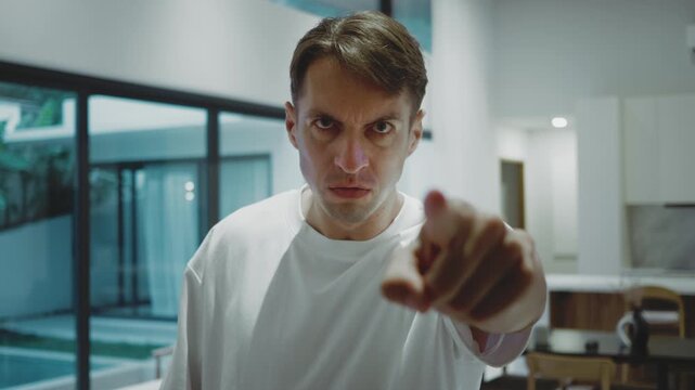 Angry man points at camera with intense stare in modern home. He points a finger and blames you, scolds you, confronts you to take responsibility and stop lies stop fraud now.