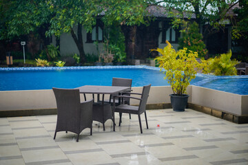 Obraz premium Outdoor patio with rattan table and chairs beside a blue swimming pool on a rainy day