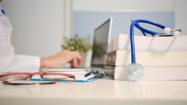 Doctor working at medical office desk, with stethoscope resting on medical textbooks and glasses on a clipboard, representing healthcare learning and professional consultation