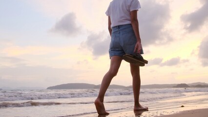 Naklejka premium Young woman walking barefoot along a sandy beach shoreline at sunset, carrying sandals in hand, enjoying freedom and peace on a mindful solo ocean stroll