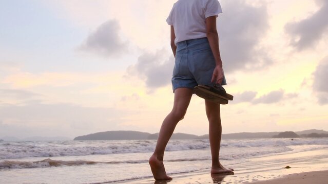 Young woman walking barefoot along a sandy beach shoreline at sunset, carrying sandals in hand, enjoying freedom and peace on a mindful solo ocean stroll