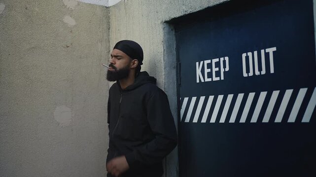 African American man in hoodie lighting cigarette at the back door of building