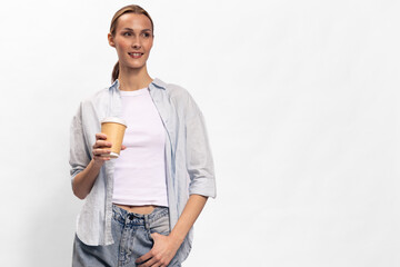 Young woman in casual clothes holding disposable coffee cup looks away with slight smile. White background, studio lighting. Concept of relaxation, lifestyle, break, and casual fashion.