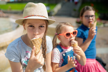 Happy group of children eating ice cream cones together by city park fountain on sunny summer day, wearing colorful casual clothes and hats