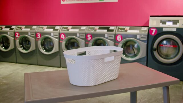 White laundry basket placed on a table in a self-service laundromat with washing machines lined up against a vibrant pink wall in the background
