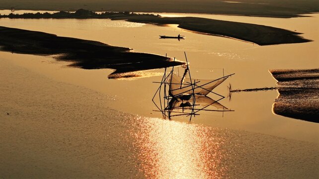 A traditional fishing net stands silhouetted against the golden waters of the Brahmaputra River at sunset in Majuli, Assam, as sandbars emerge during the dry season