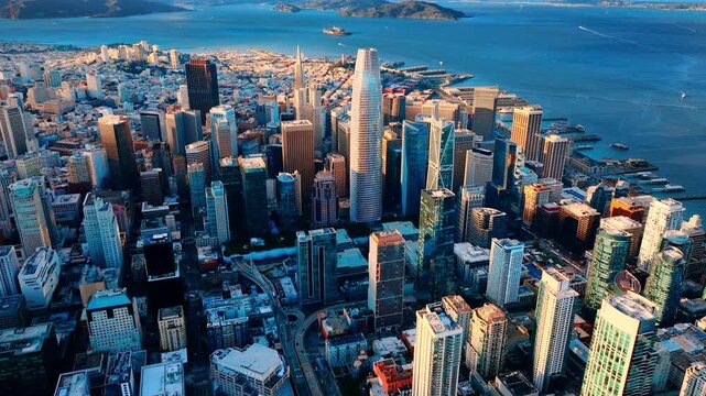 Aerial view of the San Francisco financial district during sunset. The low sun illuminates the tops of skyscrapers and the bay with blue water. Ships on the water and Alcatraz Island.