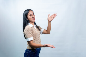 Young Southeast Asian woman in a sweater vest making a presenting gesture with both hands. Professional studio portrait on white background. 