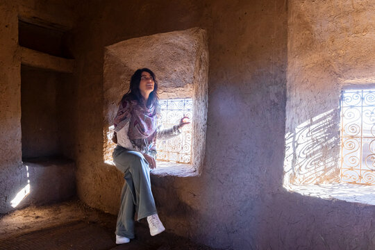 A woman sits in a window of a mud-brick building, looking up at the light. Kasbah Amridil,Marrakech,Morocco