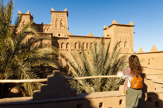 A woman with a backpack looks at the Kasbah Amridil, a traditional mud-brick fortress with palm trees, under a clear blue sky. Kasbah Amridil,Marrakech,Morocco
