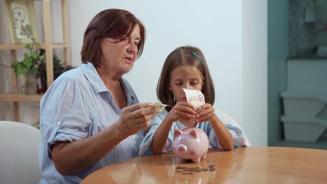 Grandmother and granddaughter inserting banknotes into piggybank while sitting at home, focusing on saving money and sharing valuable financial lessons together