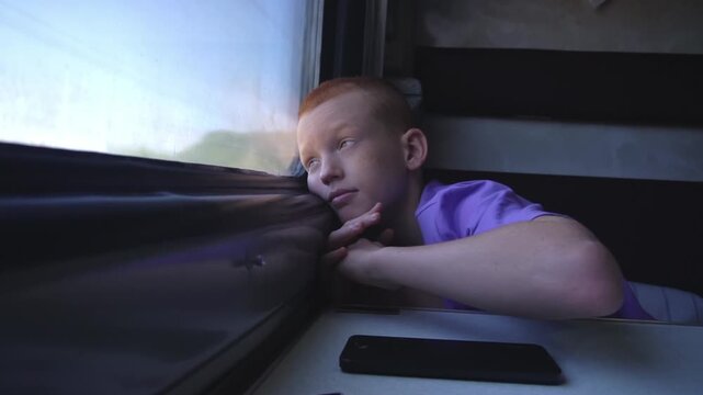 A red-haired teenager puts his phone down on the table and looks dreamily out the train window while sitting in his compartment during a journey. High quality FullHD footage