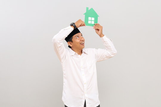 Asian Muslim man holding a small paper house image, posing in front of a gray background wall with empty space for promotional content or design.