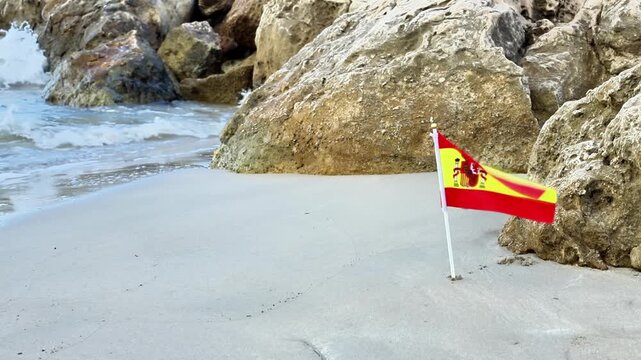Spanish flag stands on sandy beach near rocky shoreline, with gentle waves lapping at the shore, creating a serene coastal scene in a sunny environment