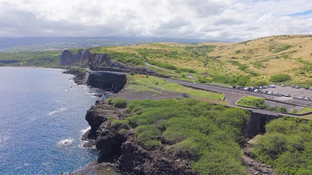Aerial drone footage with a smooth ascending movement starting near the coastline and natural pools before revealing a wider view of the coastal road and distant town in Reunion Island.